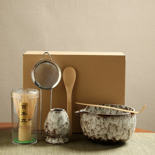 Tea-making set with a glass container, wooden spoon, and ceramic bowl on a green mat.